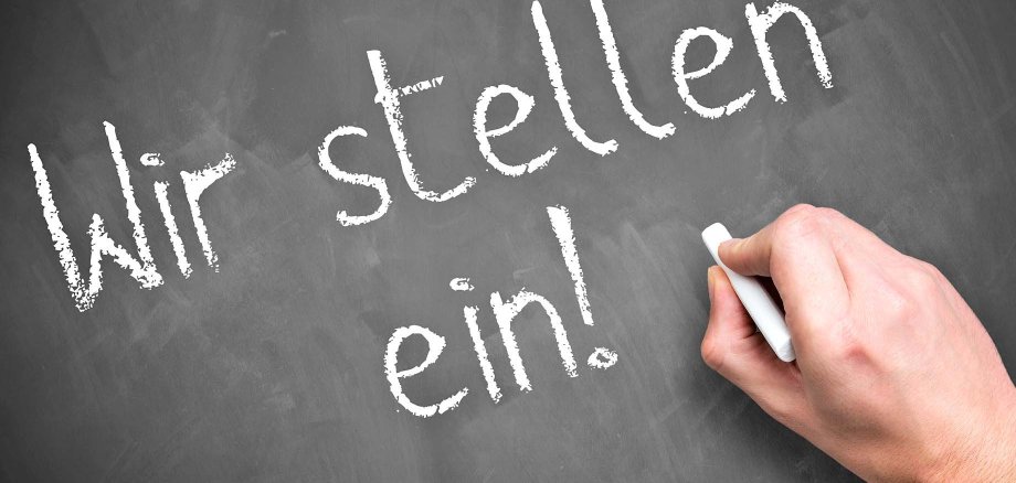 Eine Hand schreibt die Worte "Wir stellen ein!" in weißer Kreide auf eine graue Schiefertafel