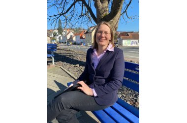 Eine blonde Frau sitzt lächelnd auf einer Bank im Freien bei Sonnenschein, im Hintergrund sind Häuser zu sehen