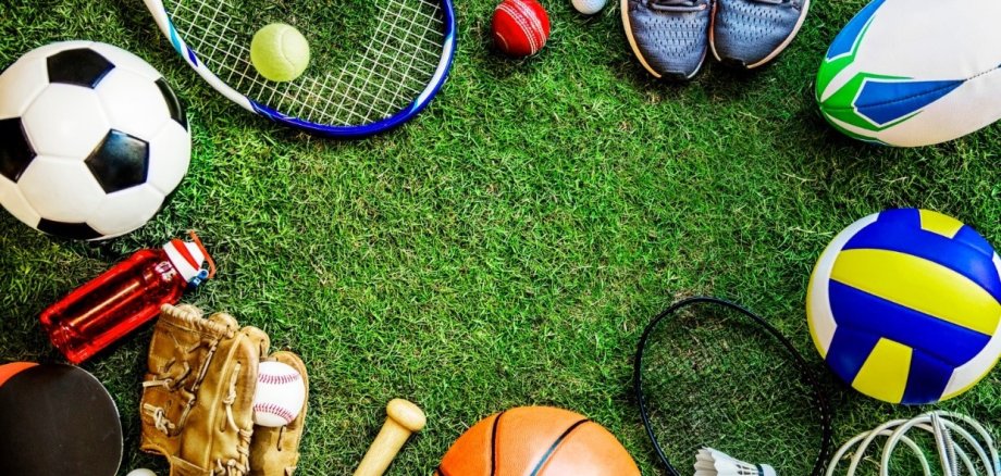 Blick von oben auf verschiedene Sportgeräte wie Bälle und Tennisschläger, die auf grünem Kunstrasen liegen