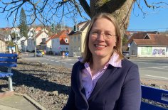 Eine blonde Frau sitzt lächelnd auf einer Bank auf einem Platz mit Häusern im Hintergrund