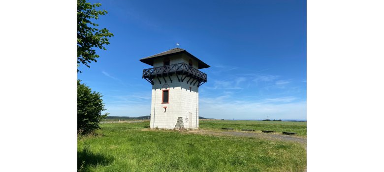 Ansicht eines weißen, quadratischen Turms mit schwarzem Schieferdach auf einer Wiese, im Hintergrund führ eine Straße vorbei