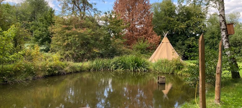 Von Bäumen umstandener Teich mit einem Tipi im Hintergrund