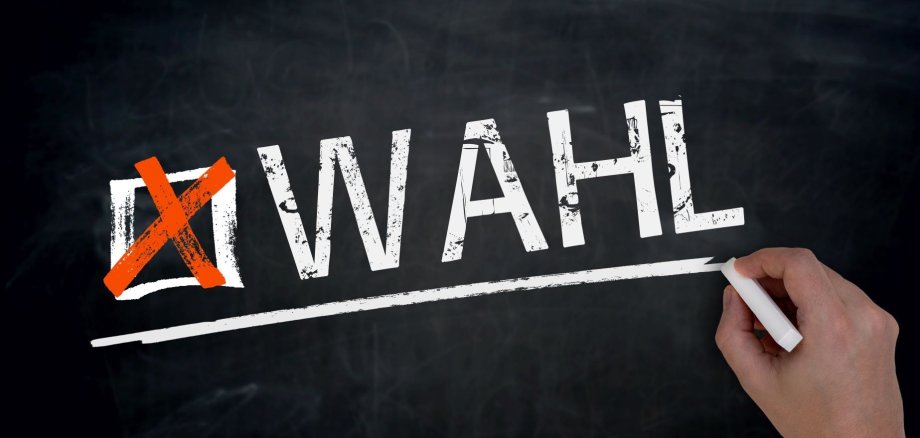 Eine Hand schreibt mit Kreide das Wort "Wahl" an eine dunkelgraue Schiefertafel