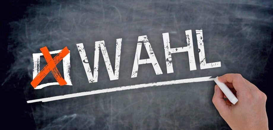 Eine Hand schreibt das Wort "Wahl" mit Kreide an eine graue Tafel