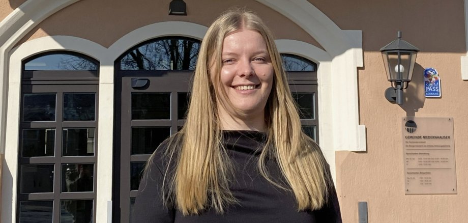 Eine junge Frau mit langen, blonden Haaren und einem schwarzen Pullover steht vor einem Gebäude, auf dessen Fassade "Rathaus" steht