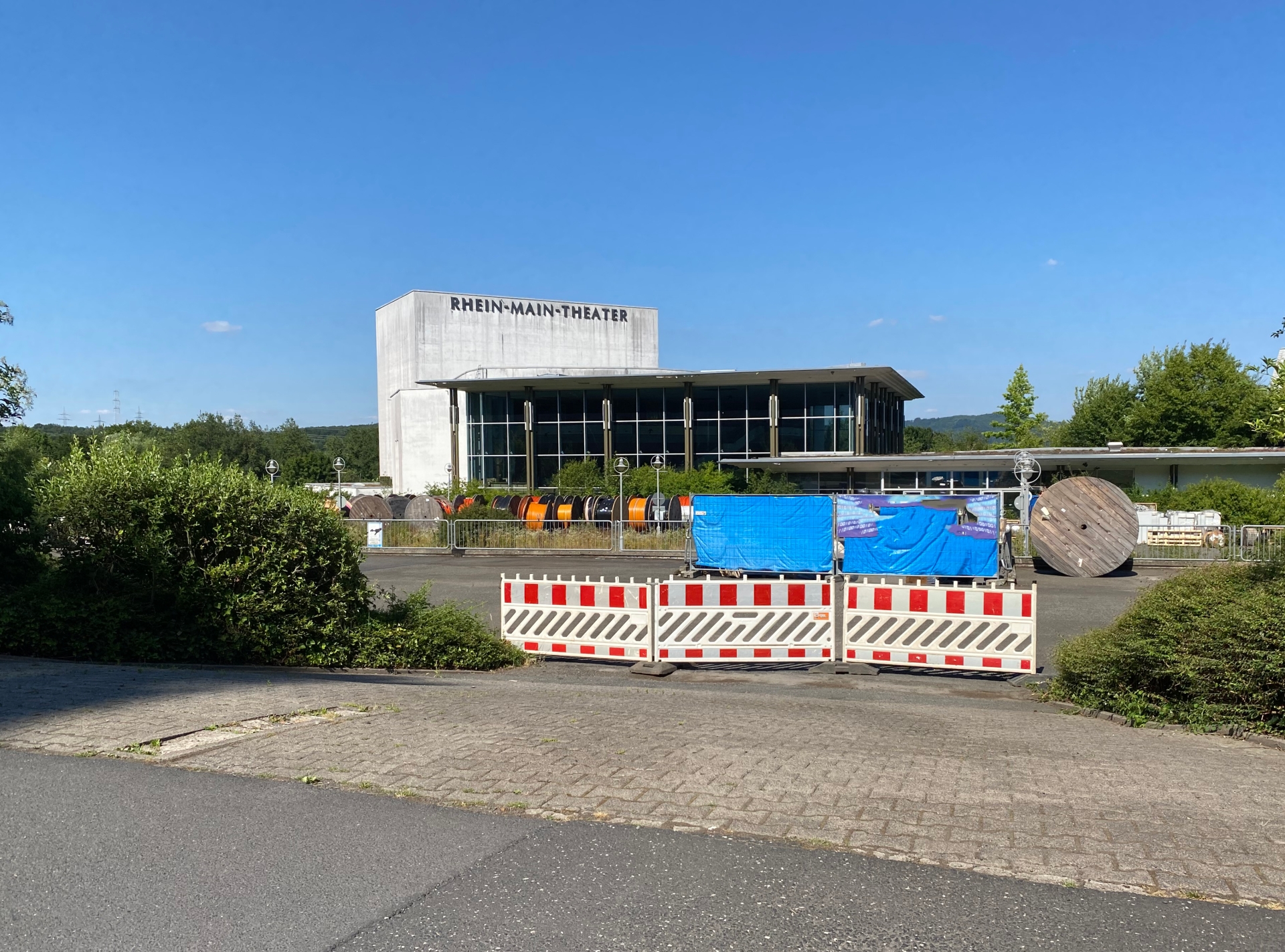 Blick auf ein hohes Gebäude aus Beton und Glas mit der Aufschrift "Rhein Main Theater", das von Bauzäunen umgeben ist