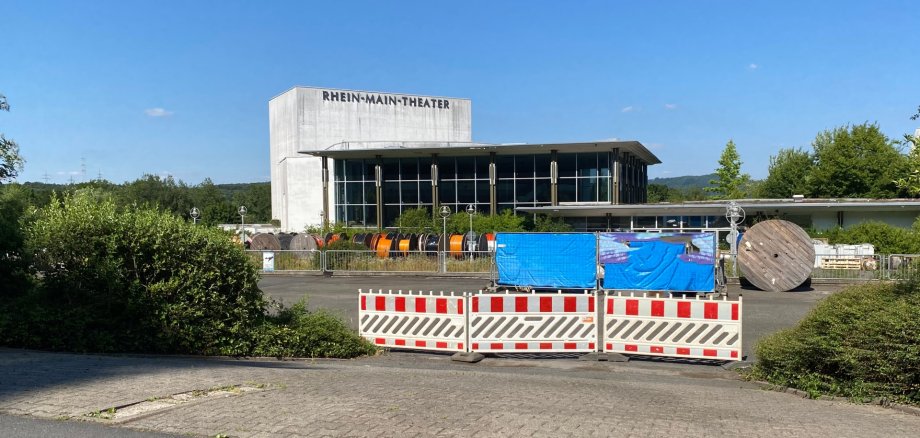 Blick auf ein hohes Gebäude aus Beton und Glas mit der Aufschrift "Rhein Main Theater", das von Bauzäunen umgeben ist