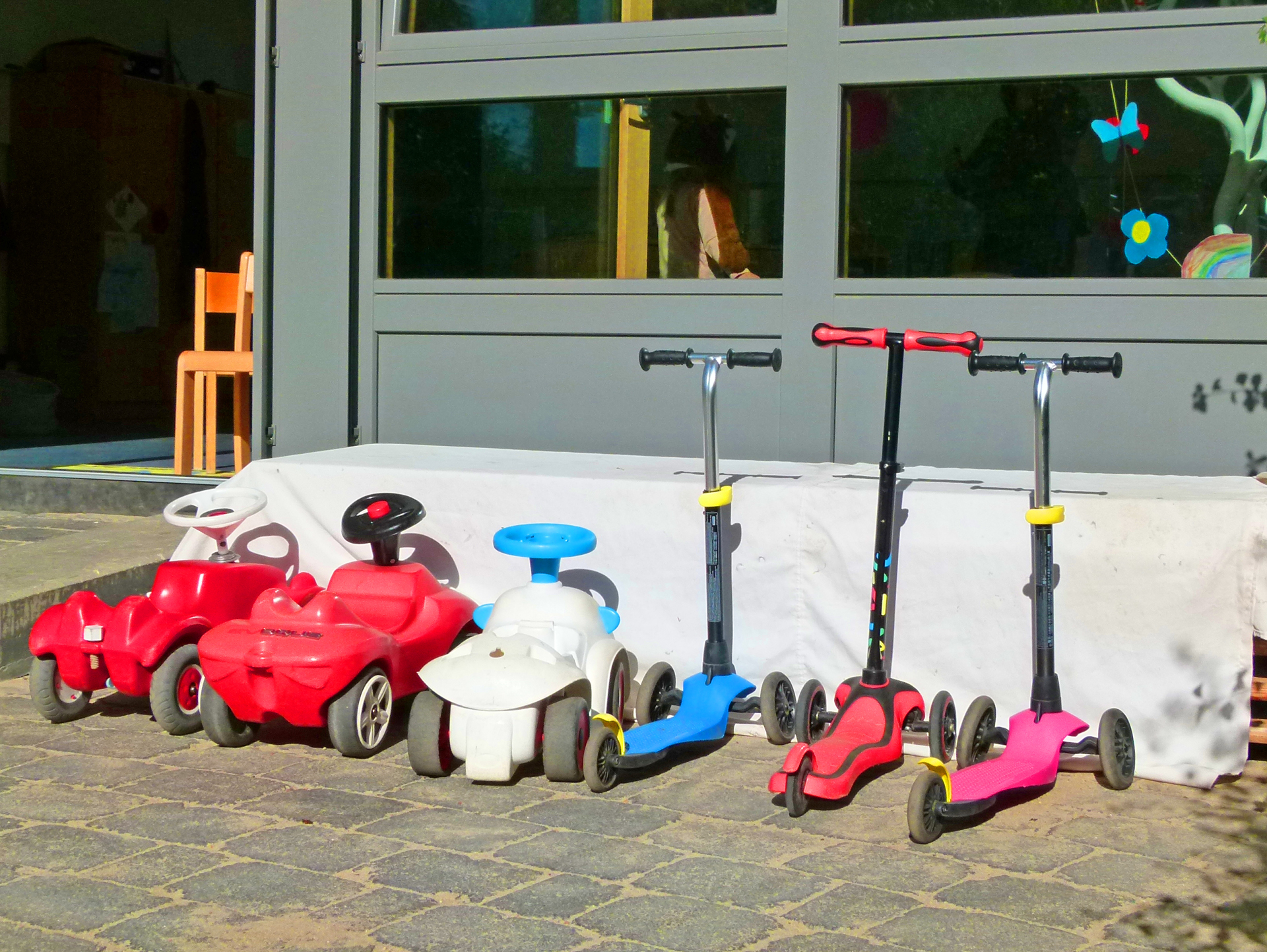 Zwei rote Bobbycars und drei bunte Roller parken im Sonnenschein vor einer gläsernen Fensterwand