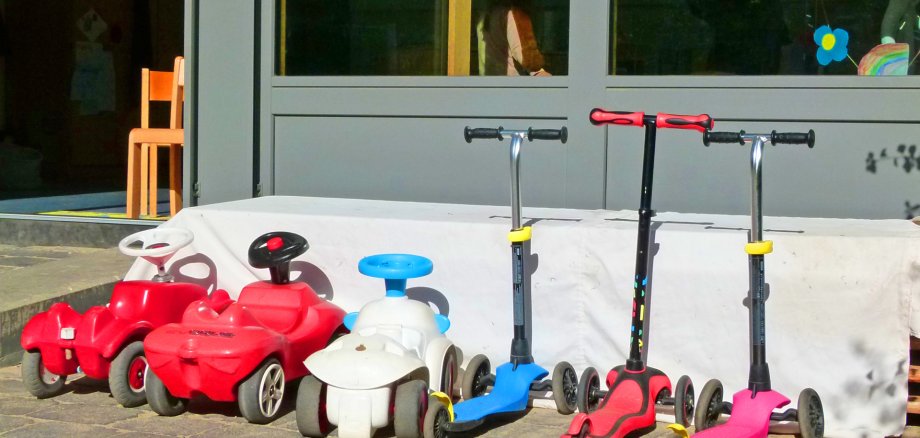 Zwei rote Bobbycars und drei bunte Roller parken im Sonnenschein vor einer gläsernen Fensterwand