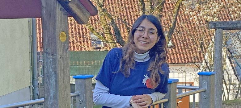 Eine junge Frau mit lockigen dunklen Haaren und Brille sitzt lächelnd auf einem Spielplatz-Klettergerüst