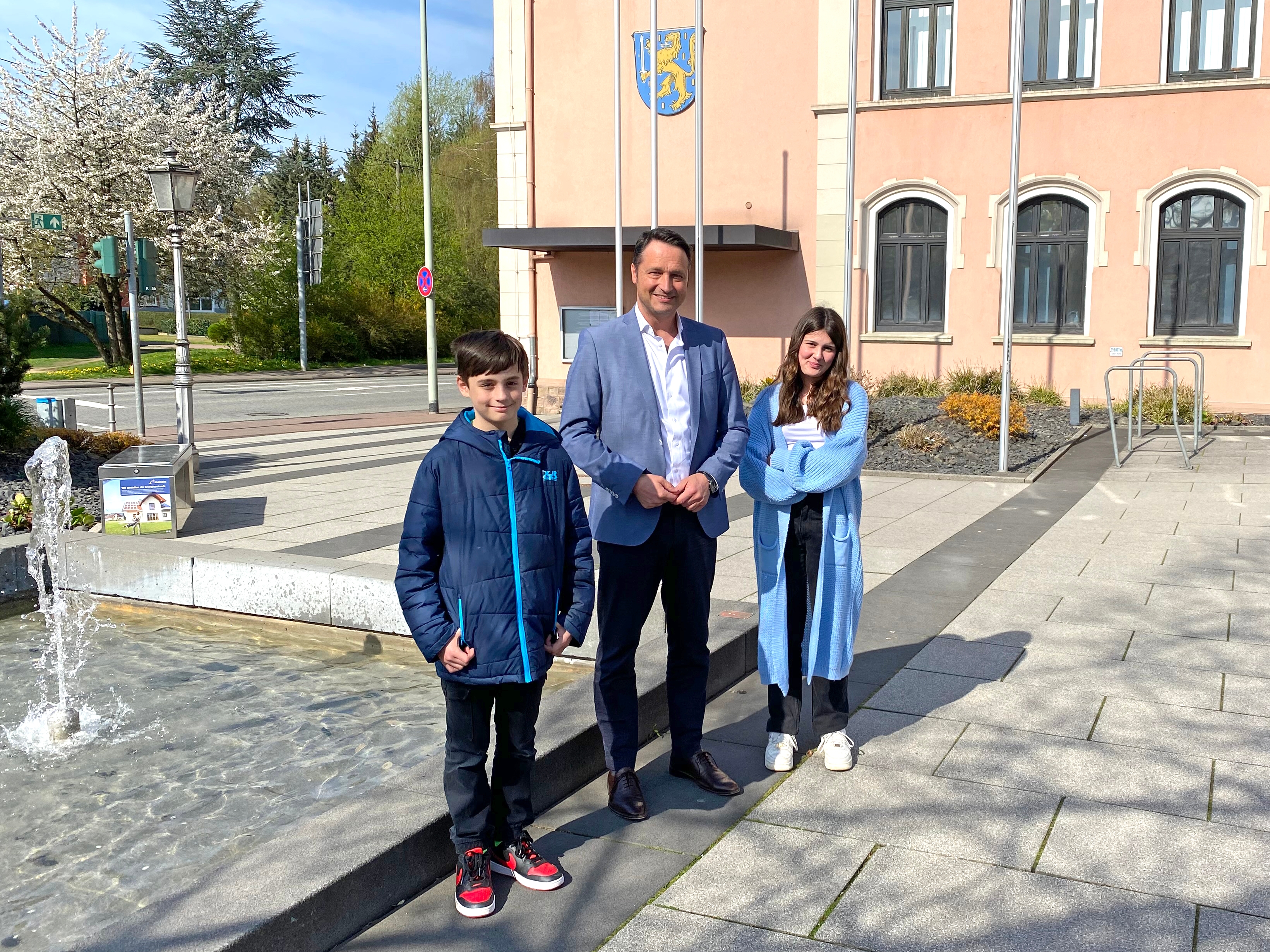 Ein Erwachsener und zwei Jugendliche stehen auf einem Platz im Freien. Im Hintergrund sieht man ein Gebäude und einen Springbrunnen.