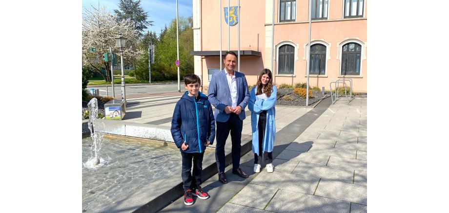 Ein Erwachsener und zwei Jugendliche stehen auf einem Platz im Freien. Im Hintergrund sieht man ein Gebäude und einen Springbrunnen.